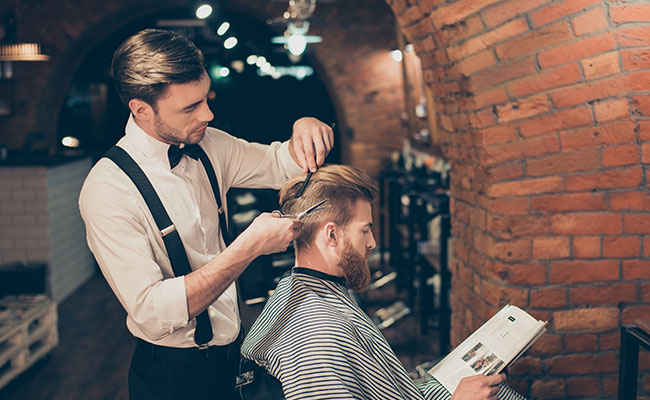 Man cutting another man's hair while he reads a magazine
